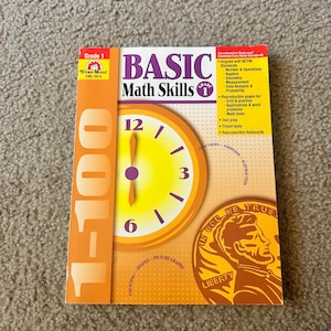 May include: A Grade 1 "Basic Math Skills" workbook by Evan-Moor. The cover features a large clock graphic with the numbers 12, 3, 6, and 9. The book includes sections on counting, shapes, and picture graphs.
