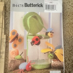 May include: A sewing pattern for a soft toy storage bag with a green felt lid and a mesh body. The pattern includes instructions for making five different felt toys: a ladybug, a snail, a butterfly, a bee, and a spider. The pattern is titled "B4478 Butterick Soft Stuff".