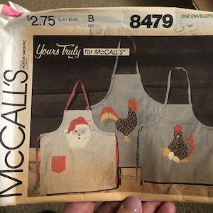 May include: A sewing pattern for three aprons with different designs. The aprons are striped and feature a Santa Claus, a rooster, and a hen with eggs. The pattern is called "Yours Truly for McCall's" and the pattern number is 8479.