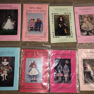 May include: A collection of eight doll-making patterns featuring various dolls, including a tea cozy, a rag doll, and a froggy-themed doll. The patterns are printed on pink and blue paper and are packaged in clear plastic bags.