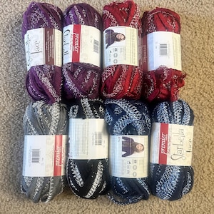 May include: An overhead shot of nine skeins of yarn in various colors, including purple, red, gray, black, and navy blue. The yarn has a metallic, lace-like texture. Each skein has a label with the brand name and product information.