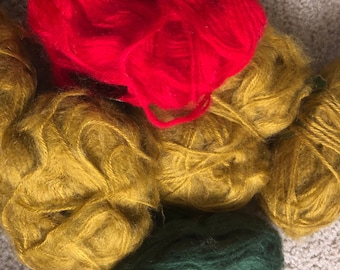 May include: Three skeins of yarn in red, gold, and green. The red yarn is in the center of the image, the gold yarn is to the left of the red yarn, and the green yarn is to the right of the red yarn.