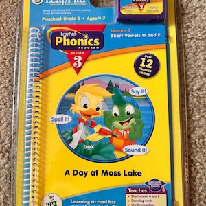 May include: A LeapPad Phonics Program Lesson 3 set, featuring a book and cartridge. The cover is yellow and blue, with cartoon characters and the text "A Day at Moss Lake." It teaches short vowels O and E for ages 4-7.