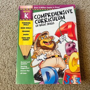 May include: A colorful Grade K workbook titled "Comprehensive Curriculum of Basic Skills." The cover features a cartoon lion holding a large red letter "A" and other letters. The book includes sections on thinking, reading, math, and time and money.