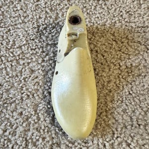 May include: A vintage, off-white shoe last, a form used to shape shoes during manufacturing. The last is made of a durable material and features a rounded toe and a heel area with a hole. The surface shows signs of wear, indicating its use in shoemaking.