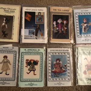 May include: Eight different doll-making patterns featuring whimsical characters. The patterns are printed on white paper with colorful illustrations of the dolls. The titles of the patterns include "Maggie", "Paper Nella", "Agnes the Flag Keeper", "Down Hill Samba", "Pearl Diver", "The Tea Ladies", "Tilly Teapot", and "Elinor Peace Bailey".