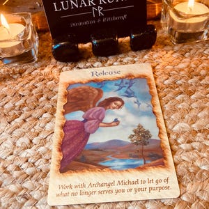 Puede incluir: Una carta de or&aacute;culo con la palabra "Release" y el texto "Work with Archangel Michael to let go of what no longer serves you or your purpose." La carta est&aacute; sobre una superficie tejida, con velas y cristales en el fondo. La carta presenta un &aacute;ngel con alas marrones.