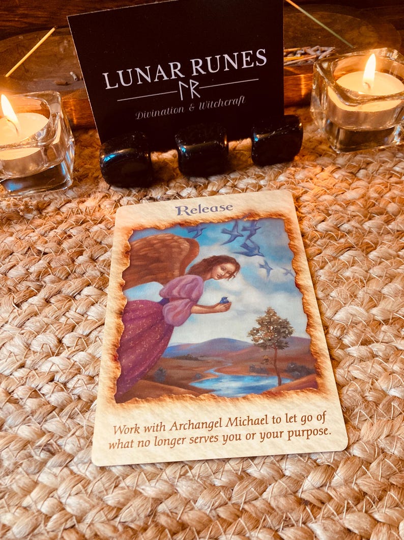 Puede incluir: Una carta de or&aacute;culo con la palabra "Release" y el texto "Work with Archangel Michael to let go of what no longer serves you or your purpose." La carta est&aacute; sobre una superficie tejida, con velas y cristales en el fondo. La carta presenta un &aacute;ngel con alas marrones.