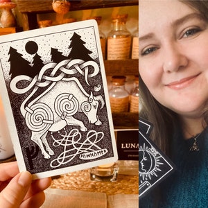 May include: A black and white tarot card featuring a bull illustration and the word "Annanann" in a stylised font. The card is held in a hand against a backdrop of wooden shelves with bottles.