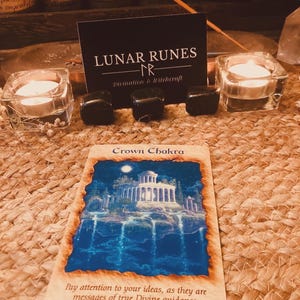 Puede incluir: Una imagen de una carta de tarot con una ilustraci&oacute;n azul y blanca de un edificio, con las palabras "Crown Chakra". La carta est&aacute; rodeada de piedras negras, velas encendidas y una tarjeta de visita "Lunar Runes".