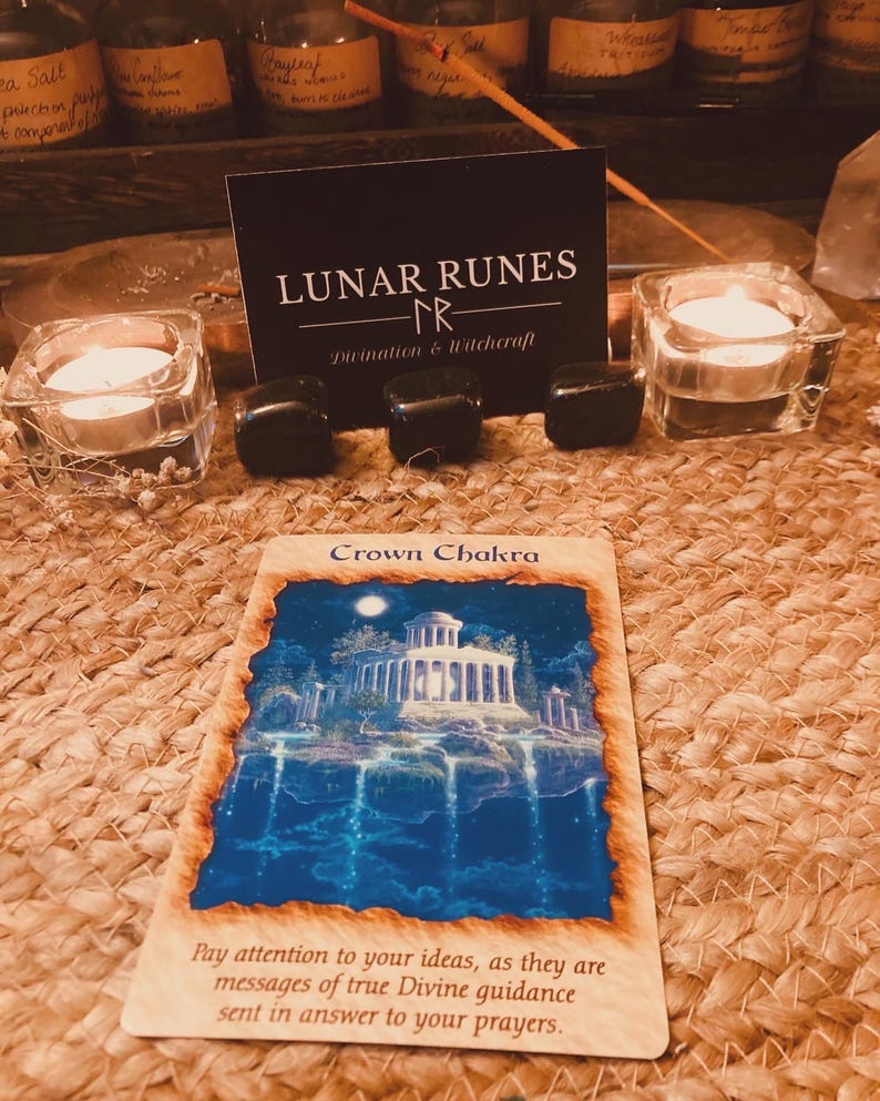 Puede incluir: Una imagen de una carta de tarot con una ilustraci&oacute;n azul y blanca de un edificio, con las palabras "Crown Chakra". La carta est&aacute; rodeada de piedras negras, velas encendidas y una tarjeta de visita "Lunar Runes".