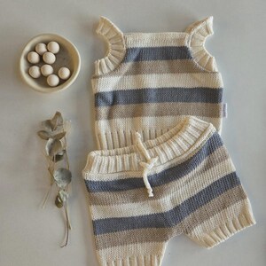 May include: A knitted baby outfit in cream, gray, and tan horizontal stripes. The set includes a sleeveless top with ruffled shoulders and matching shorts with a drawstring. A small bowl with white spheres and dried foliage are also in the image.