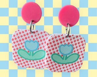 May include: A pair of pink and white square earrings with a blue flower design. The earrings have a pink circle at the top and a silver hook.