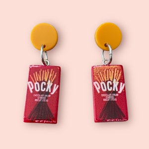 Peut inclure: Une paire de boucles d'oreilles avec un charme en forme de boîte Pocky rouge et or avec le texte "Pocky Chocolate Cream Covered Biscuit Sticks" et un charme rond jaune.