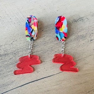 May include: A pair of dangle earrings with a red, blue and yellow abstract pattern on the top and a red, wavy, abstract shape on the bottom.