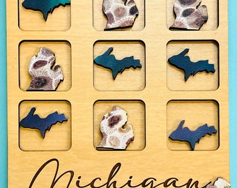 Puede incluir: Un juego de tres en raya de madera con el contorno del estado de Michigan. Las piezas del juego están hechas de madera y presentan diferentes diseños, incluyendo un degradado azul y verde, un patrón floral marrón y blanco y un color azul sólido. El tablero de juego está etiquetado como "Michigan Tic-Tac-Toe".