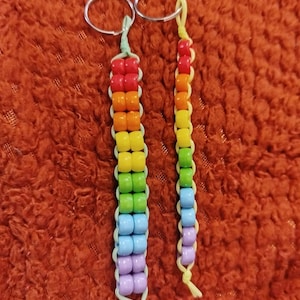 Puede incluir: Dos llaveros hechos a mano con cuentas de colores del arcoíris. Los llaveros presentan una serie de cuentas coloridas en un patrón de arcoíris, unidas a un anillo de metal plateado. Las cuentas son rojas, naranjas, amarillas, verdes, azules y moradas. Los llaveros están sobre un fondo naranja.