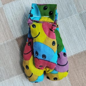 Puede incluir: Un artículo de tela colorido y hecho a mano con un patrón repetido de caras sonrientes en varios colores, incluyendo amarillo, azul, rojo y morado. El artículo parece estar doblado, con una costura visible y un cierre de botón. El fondo es una superficie neutra estampada.