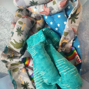 May include: A collection of fabric strips featuring various patterns including dinosaurs on a light grey background, white stars on a blue background, a solid teal textured fabric, and a white fabric with a rainbow and the word love printed on it.