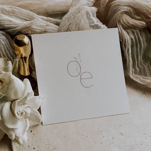 May include: A square, off-white card with the letters "oe" in a delicate, minimalist design. A gold-colored bottle stopper and a white flower are visible. Soft, neutral-toned fabric drapes across the surface, creating a serene and elegant aesthetic.