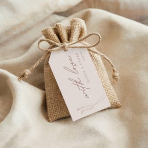 Pode incluir: Uma pequena bolsa de serapilheira amarrada com barbante, com uma etiqueta retangular. A etiqueta diz "with love" em escrita elegante, junto com nomes e uma data. A bolsa e a etiqueta são marrons claras, em um fundo neutro.