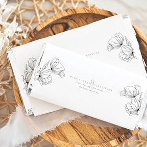 Pode incluir: Embalagens de barras de chocolate brancas com arte de linha floral preta e o texto "WITH LOVE and GRATITUDE". As embalagens estão em uma bandeja de madeira com um fundo tecido e flores secas.
