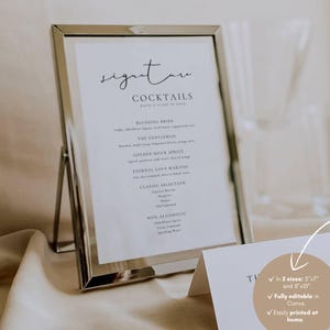 Pode incluir: Uma placa com moldura prateada com as palavras "Signature Cocktails" em escrita elegante. A placa lista nomes e ingredientes de coquetéis, incluindo "Blushing Bride" e "Eternal Love Martini". Um cartão menor com texto está em primeiro plano.