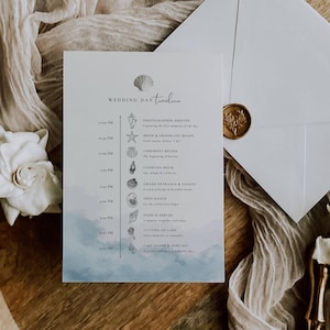 Può includere: Una scheda della cronologia del giorno del matrimonio con una grafica a conchiglia in alto. La scheda ha un design ad acquerello blu e bianco nella parte inferiore. Una busta con un sigillo d'oro è accanto alla scheda.