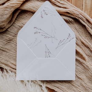 May include: A white envelope with a floral design on the inside, set on a wooden surface with a tan fabric. The envelope's interior has a delicate pattern of small flowers and stems in a soft, muted colour.