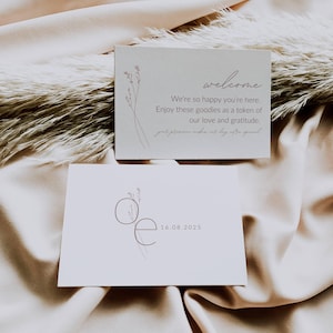 Pode incluir: Dois cartões de boas-vindas creme com delicados desenhos florais. O cartão superior diz "welcome" e uma mensagem de gratidão. O cartão inferior apresenta as iniciais "Oe" e a data 16.08.2025. Os cartões repousam sobre um tecido bege macio.