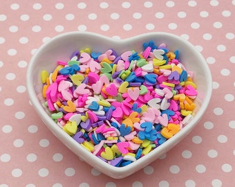 Op de afbeelding: Een witte hartvormige kom gevuld met kleurrijke hartvormige sprinkles. De sprinkles zijn in verschillende tinten roze, paars, blauw, geel en groen.