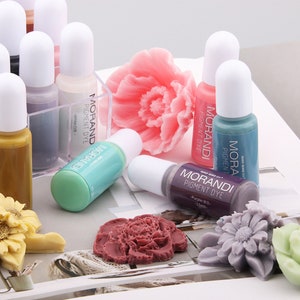 May include: Assortment of small bottles of paint pigment dye in various colors, including white, purple, teal, and blue. The bottles are labeled "Morandi Pigment Dye" and have a white cap. The bottles are arranged on a table with a variety of flowers and other decorative items.