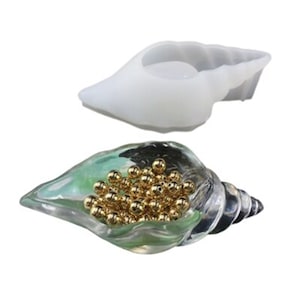 May include: A clear seashell-shaped mold with a white silicone seashell mold on top. The clear seashell is filled with gold beads.