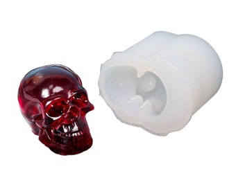 May include: A translucent, dark red skull figurine next to a white silicone mold. The skull has detailed features, including eye sockets, a nose cavity, and teeth. The mold is cylindrical with a skull-shaped cavity.