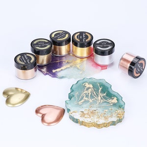 May include: Six small jars of metallic powder in various colors, including gold, silver, copper, and rose gold. The jars are arranged on a white surface with two heart-shaped pieces of metal.