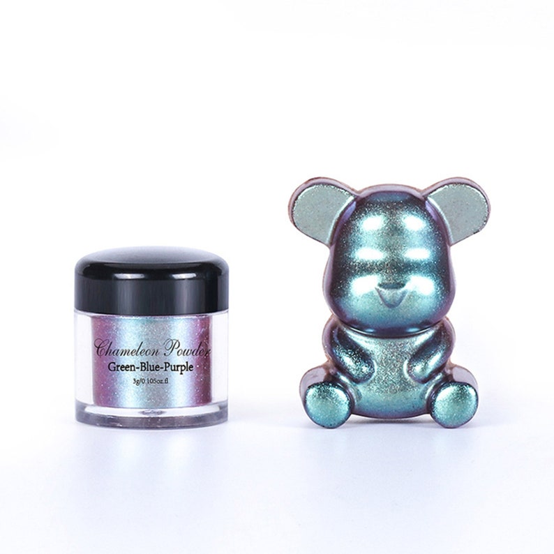 May include: A small jar of chameleon powder in green, blue, and purple shades. The jar has a black lid and a label that reads "Chameleon Powder Green-Blue-Purple 1g/0.10oz". A small, iridescent green, blue, and purple bear figurine is sitting on a white surface.