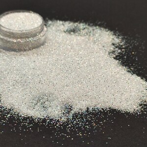 May include: A small jar of iridescent white glitter, with a portion of the glitter spilled out onto a black surface.