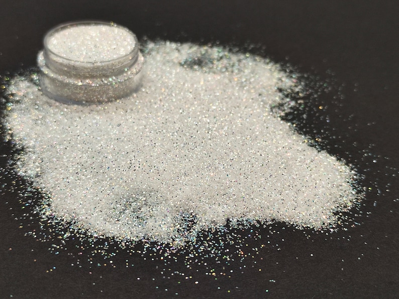 May include: A small jar of iridescent white glitter, with a portion of the glitter spilled out onto a black surface.