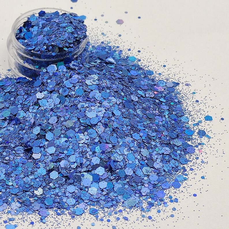 May include: A close-up of a pile of blue iridescent glitter. The glitter is scattered on a white surface.