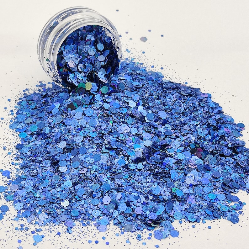 May include: A jar of blue iridescent glitter, with a variety of sizes and shapes of glitter. The glitter is spilling out of the jar onto a white surface.