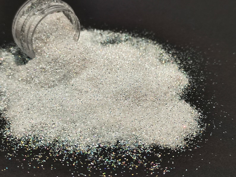May include: A pile of iridescent white glitter on a black surface. The glitter is spilling out of a clear jar.