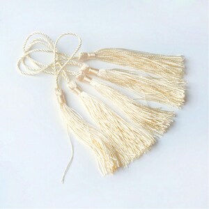 May include: Five cream-colored tassels with long, thin strands. The tassels are tied together at the top and are hanging down.