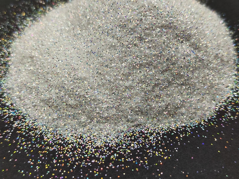 May include: A pile of iridescent white glitter with a rainbow sheen. The glitter is on a black surface.