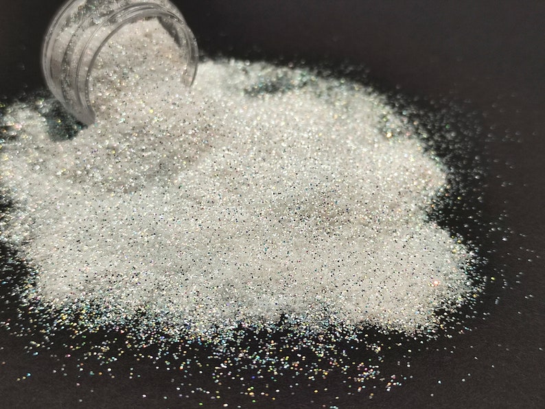 May include: A pile of iridescent white glitter, spilling out of a clear jar onto a black surface.