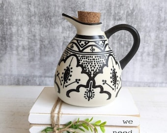 May include: White ceramic oil or vinegar bottle with a black cork stopper and black and white geometric design. The bottle is sitting on a stack of white books with the text "we need each other" printed on the books.