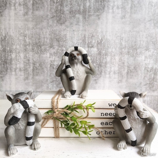 May include: Three gray and white lemur figurines with black and white striped tails covering their eyes. The lemurs are sitting on a stack of white books with a brown twine bow and a green plant.