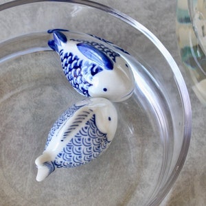 May include: Two white ceramic fish with blue patterns in a clear glass bowl filled with water. The fish are positioned one above the other. The bowl is clear, creating a decorative display. The fish are a decorative item.