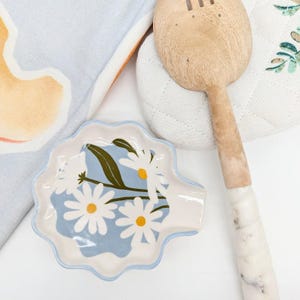 以下が含まれることがあります： 青と白の花柄の白いセラミック製のさじ置き。さじ置きは波打つ縁取りがあり、スタイリッシュなひなぎく模様が特徴です。