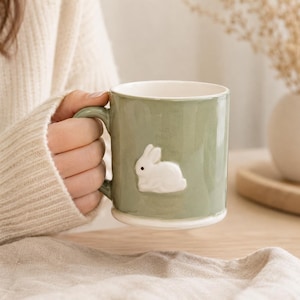 May include: A sage green ceramic mug with a white bunny relief. The mug has a rounded handle and a white interior. The mug is held by a person wearing a cream-coloured jumper. A vase with dried flowers is in the background.
