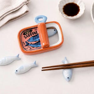 May include: A ceramic chopstick rest set featuring a rectangular orange sardine tin container with a blue fish illustration and the text SARDINE, accompanied by four small, light blue, fish-shaped ceramic rests for holding chopsticks.